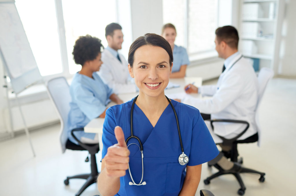 Happy Nurse giving thumbs up in front of group of medical professionals sitting at a table