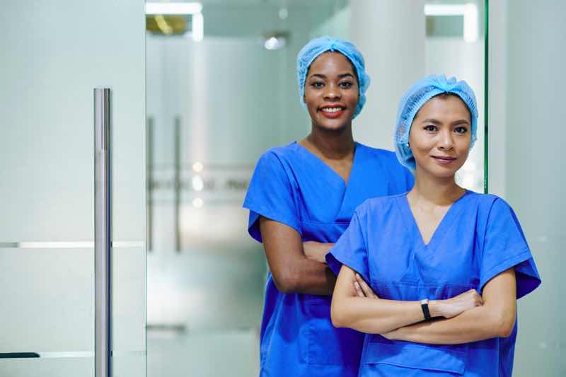 Smiling pretty multi-ethnic medical nurses in scrubs and disposable caps standing in hospital corridor